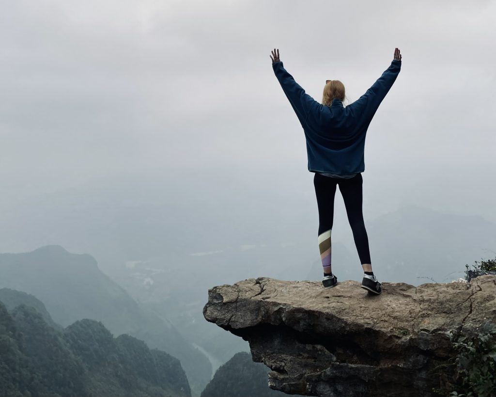 A person stands on a rock jetting out over a ledge with their arms upstretched, the background is misty Northern Vietnam scenery